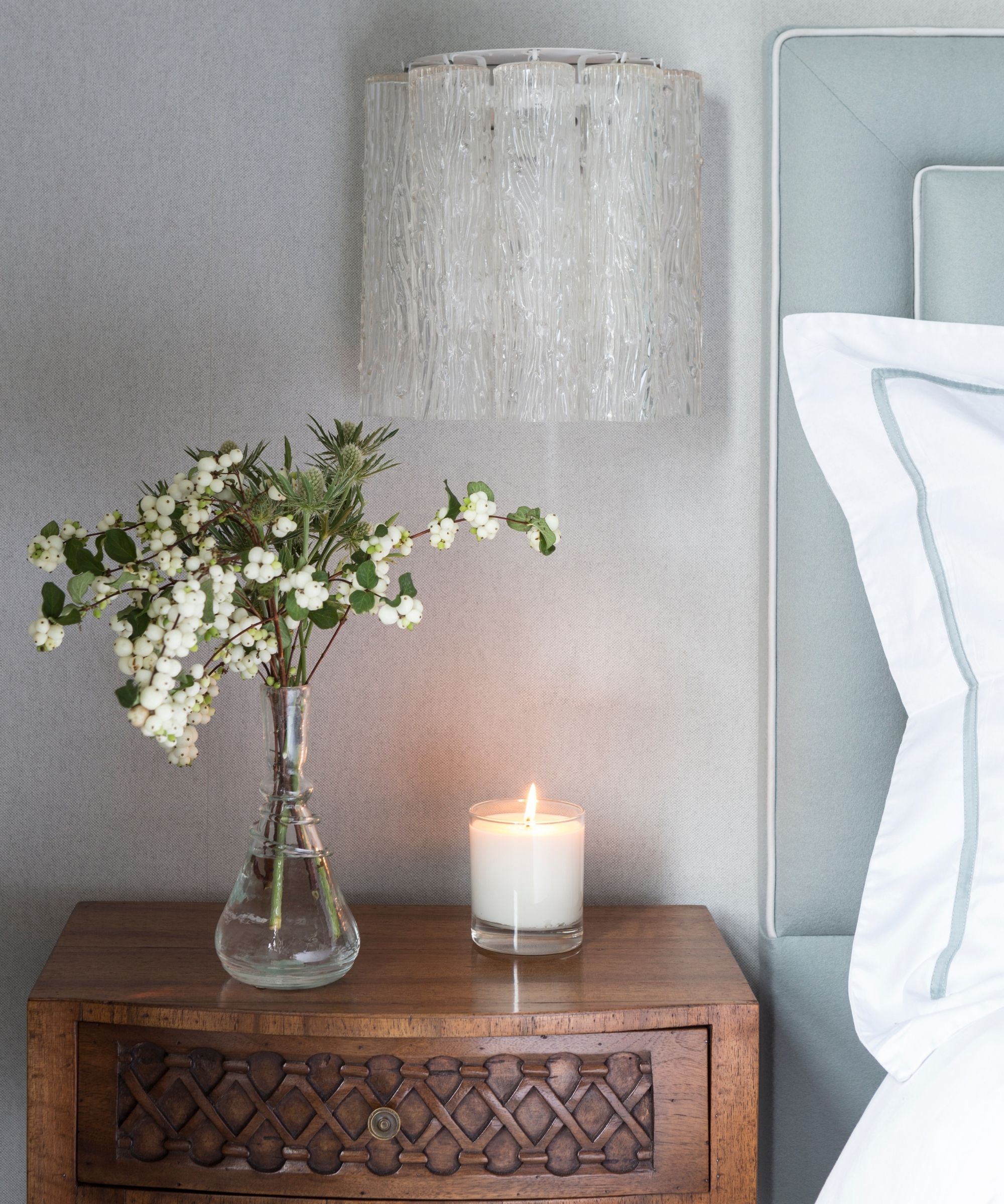Small white candle burning on a dark wood nightstand beside a glass vase of small white flowers. The bed to the right is pale blue with a white pillow. Above is a glass sconce lamp on a grey wall.