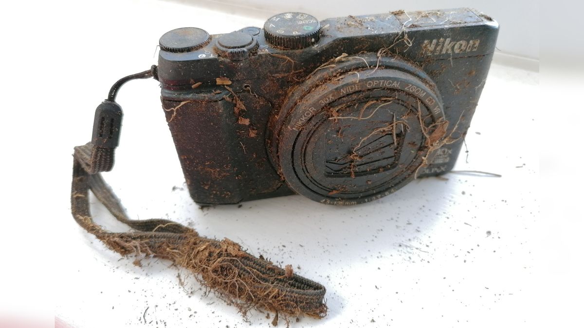 A dirty, weathered Nikon Coolpix SS9900 compact camera covered in mud and roots, resting on a white surface