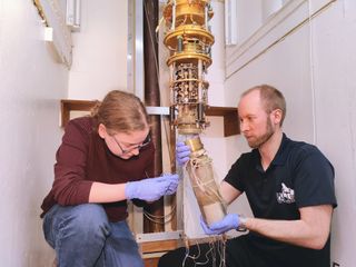 A white room containing two crouching people attending to a complex gold machine