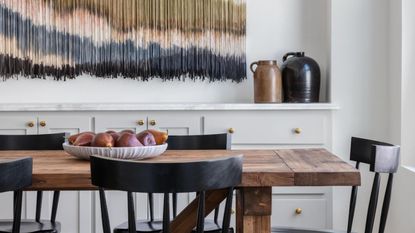 A wooden dining table with black wooden chairs. A white bowl of apples on the table. A white sideboard in the background, with a dyed string hanging on the wall above it. 
