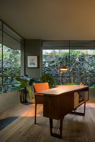 Wooden office desk with book shelf at Ventana House in Mexico, orange chair, potted plants, floor standing lamp, grey walls, wall art, windows, outside garden area