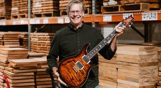 PRS Guitars COO Jack Higginbotham poses with a PRS SE Custom 24 Semi-Hollow Piezo in front of stacks and stacks of tonewood blanks.