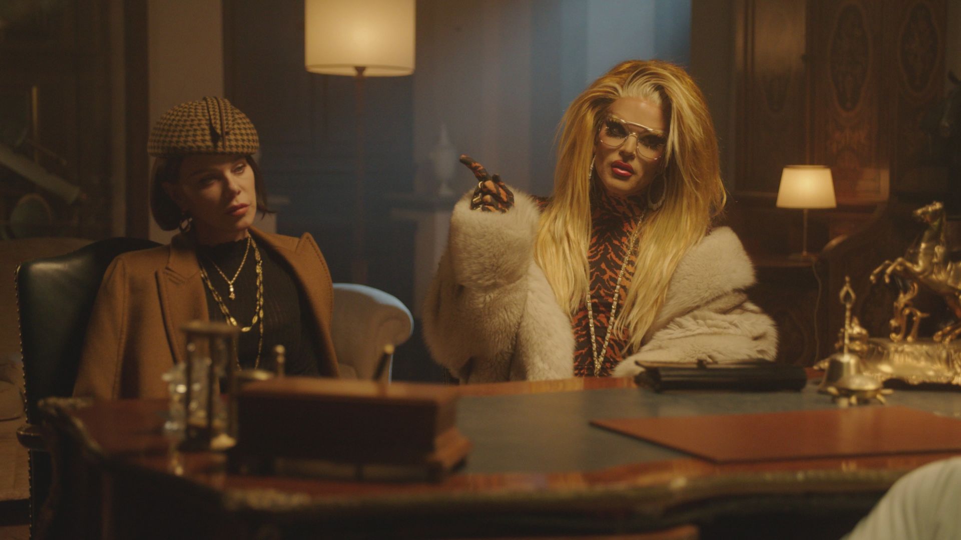 debi mazar waring a hat and blazer sitting at a table with willam belli wearing a white fur suit in a still from the mob