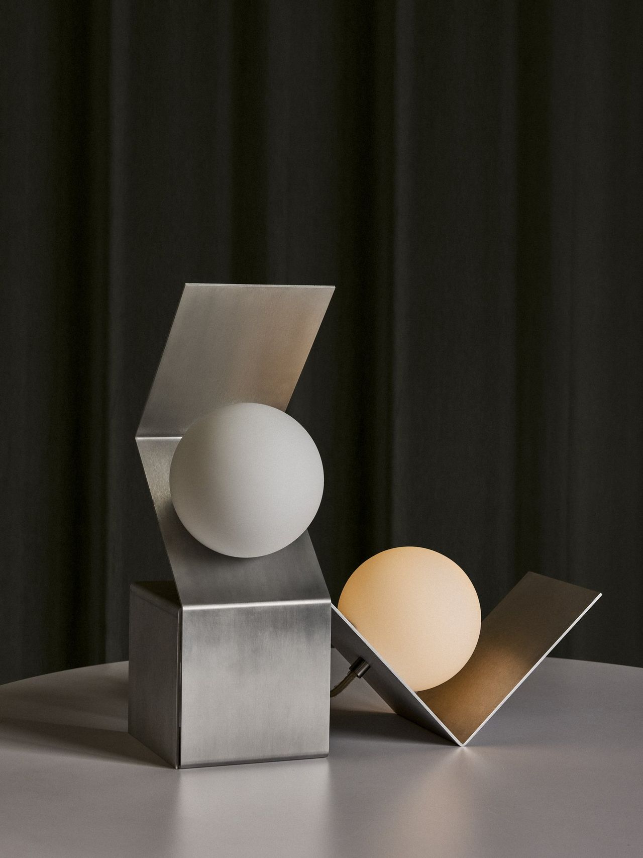 Two imaginative table lamps are composed of a silver, linear structure and a round, softly lit round light sphere.