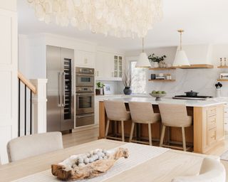 Light wood dining table with neutral upholstered chairs looking towards kitchen with neutral cabinets, wood island, neutral countertop and backsplash