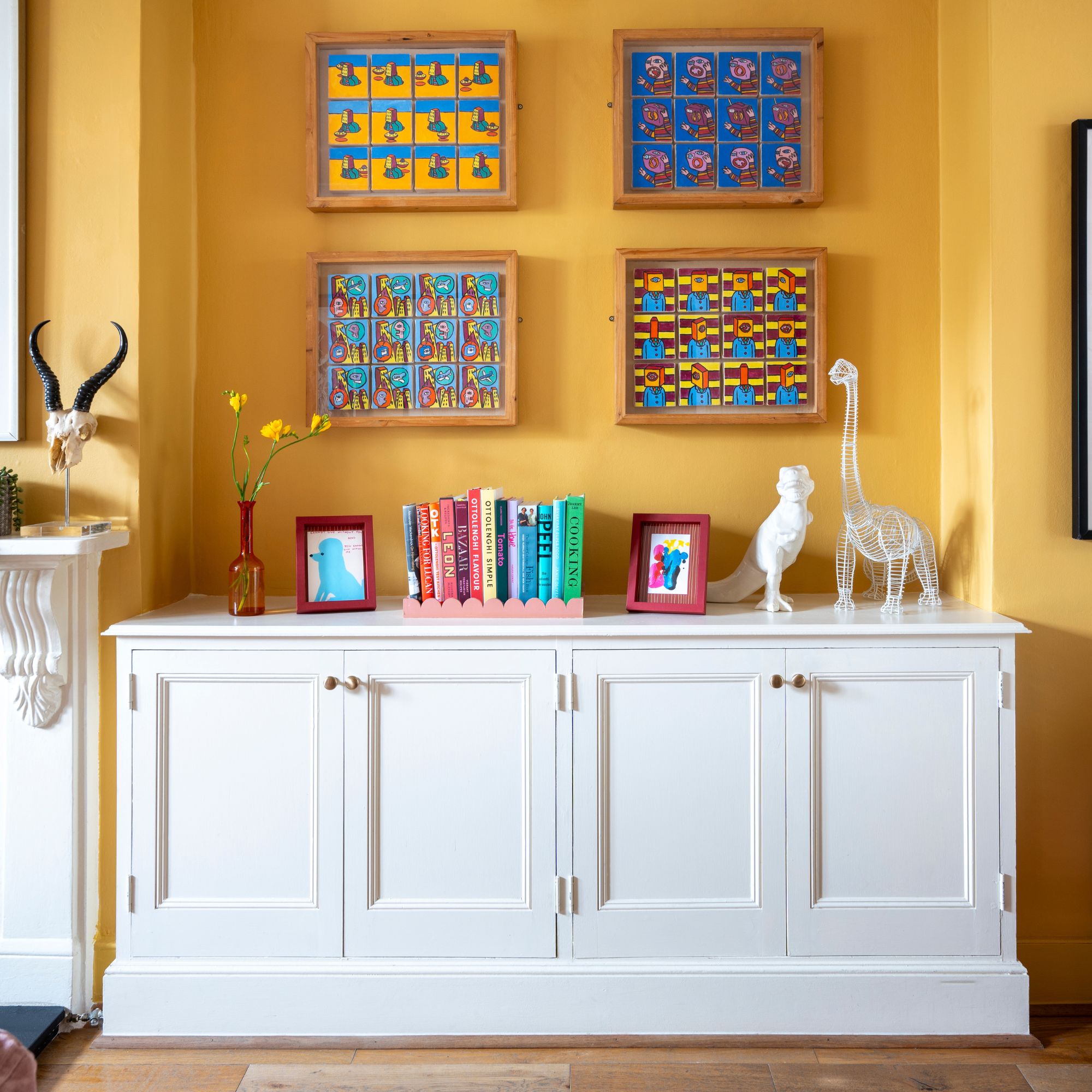 Gabinete de madera pintado de blanco cubierto de libros y baratijas frente a una pared amarilla al atardecer con obras de arte