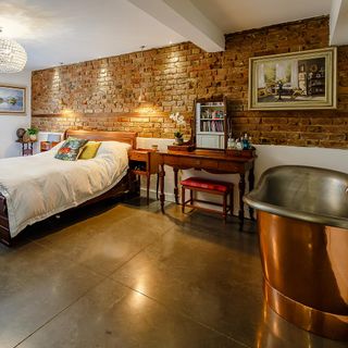 Bedroom with bed with white linen in front of exposed brick wall and a freestanding copper Victorian bath in the corner