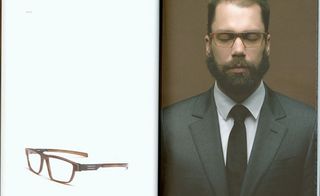 Pair of brown framed glasses on a white back ground, to the right a bearded male, smartly dressed in a grey blazer, white shirt and dark grey tie, wearing the brown framed glasses with his eyes closed, brown background