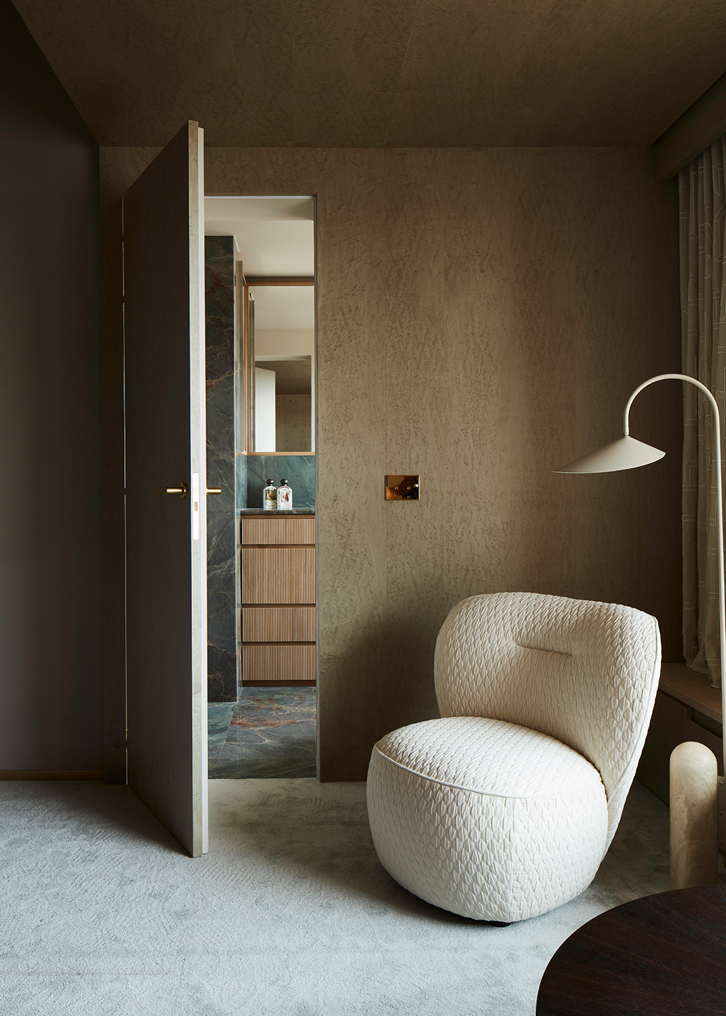 A chubby, textured armchair sits next to a standing lamp with a minimal design, colored in beige, in an earthily tinted bedroom.