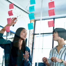 Two women discuss work in a conference room, using note cards and a clear glass board. 