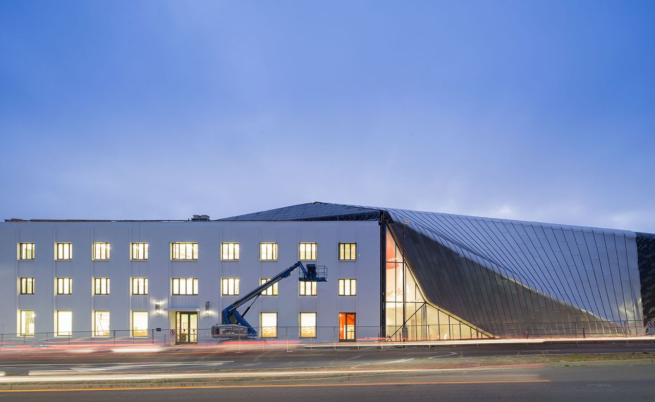 A street level view of the new museum from outside. The rectangular building has three floors with 9 columns of windows that are illuminated. To the right the building continues back with an arch-style roof. 