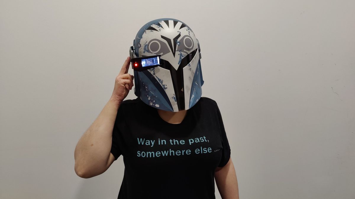 Person wearing the Black Series Bo-Katan helmet with the heads-up display down and lit up. They have their finger on the side of the helmet to make it look like they&rsquo;re using the heads-up display. They are wearing a black t-shirt with light blue text that reads &ldquo;Way in the past, somewhere else&hellip;&rdquo;.
