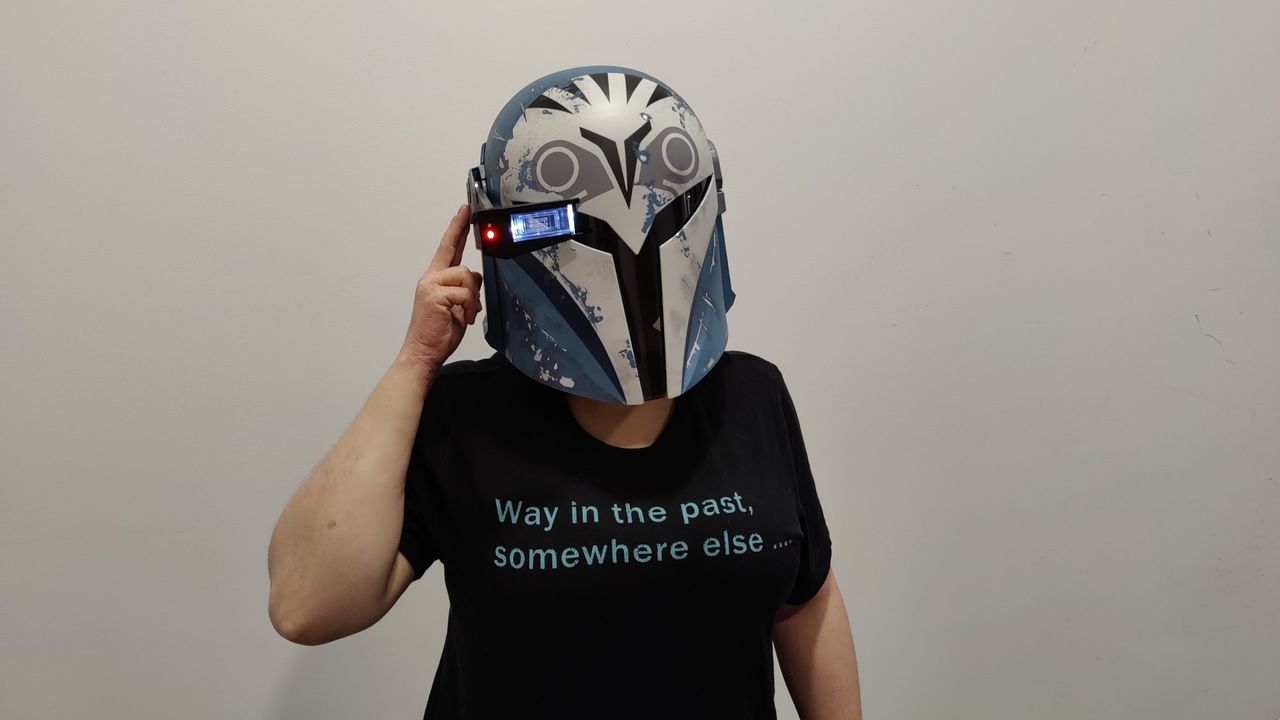 Person wearing the Black Series Bo-Katan helmet with the heads-up display down and lit up. They have their finger on the side of the helmet to make it look like they&rsquo;re using the heads-up display. They are wearing a black t-shirt with light blue text that reads &ldquo;Way in the past, somewhere else&hellip;&rdquo;.