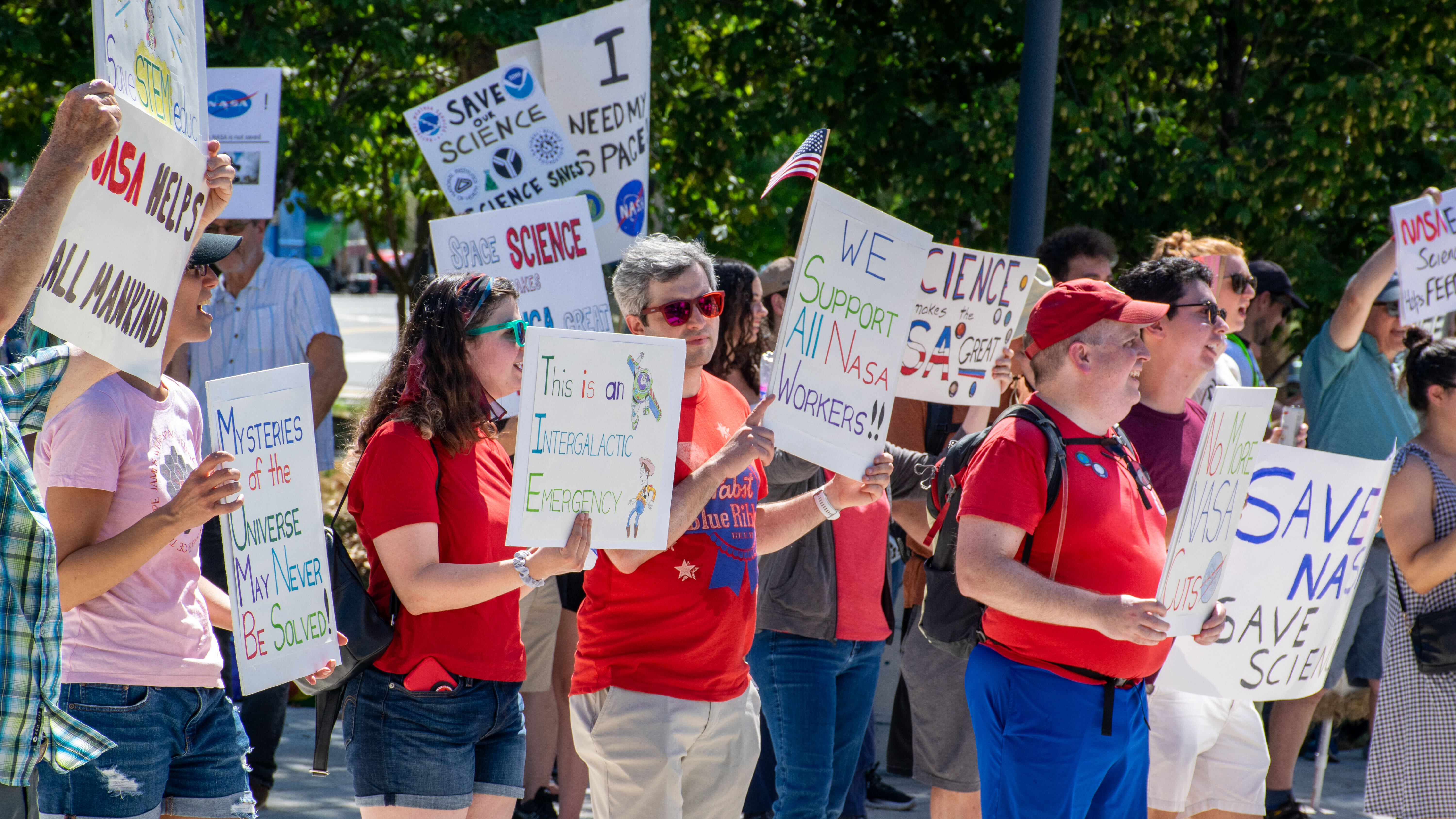 People hold signs outside on a sunny day in protest of NASA budget cuts, July 20, 2025.