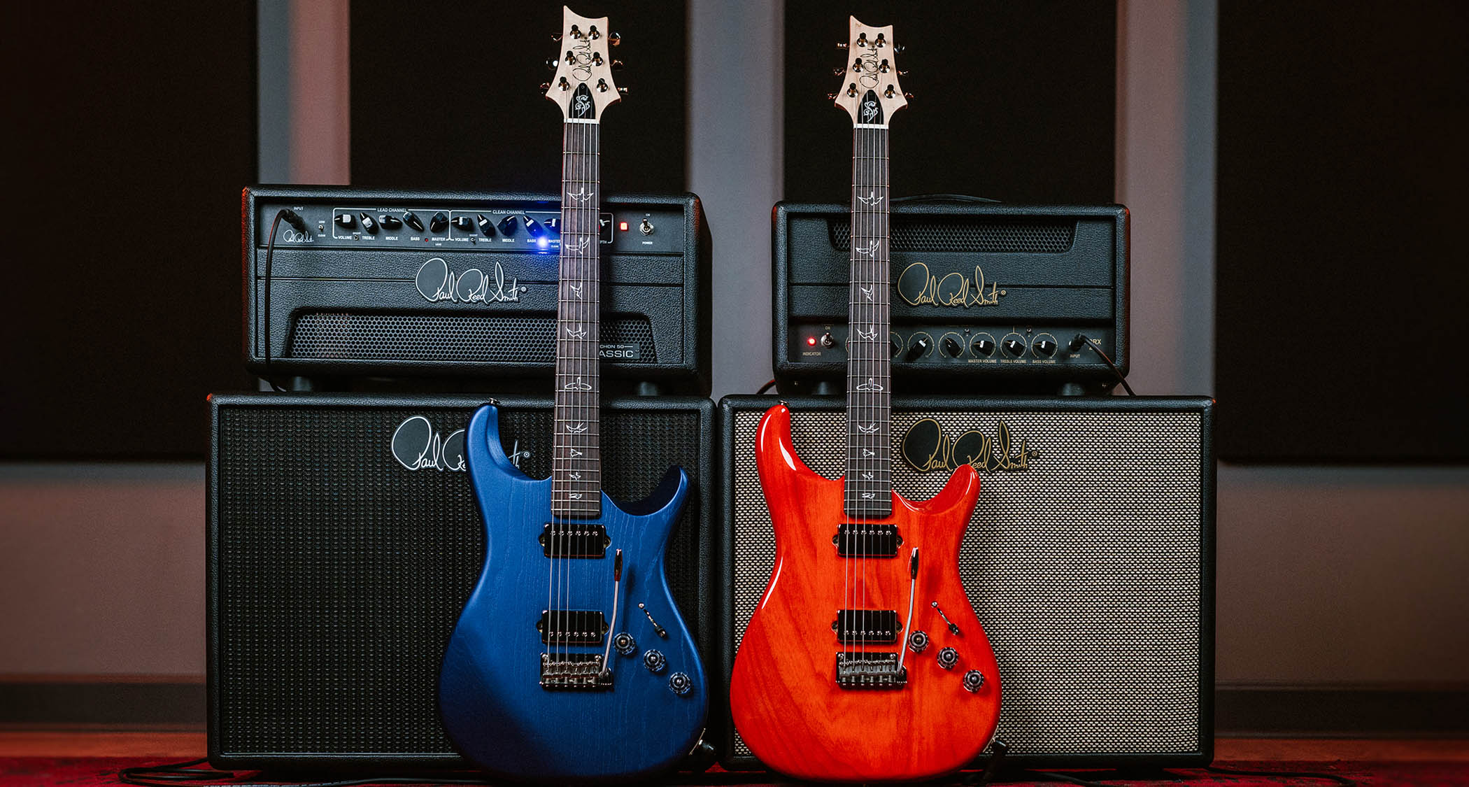 PRS Mark Lettieri Fiore HH, pictured here in its blue gloss and red satin versions against a pair of PRS tube amp stacks.