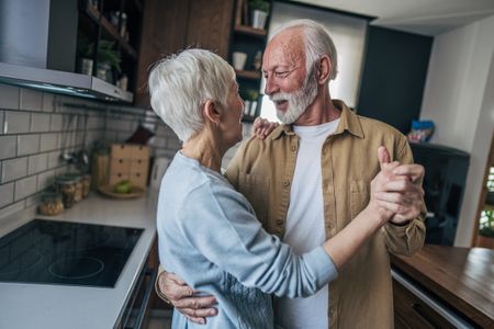 A senior couple dancing together in their kitchen. The senior man and woman are enjoying a joyful moment, holding each other as they move rhythmically to music. They smiling, sharing a special connection, and the scene is filled with warmth and happiness.