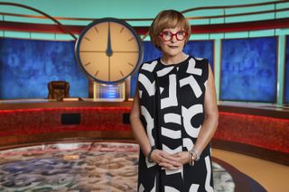 Anne Robinson in a patterned black and white dress and red-rimmed glasses, standing in the Countdown studio in front of the show's famous clock