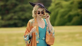 Joanna Vanderham in a blue dress and orange jacket with a purple hat and sunglasses as Diana Mitford in Outrageous