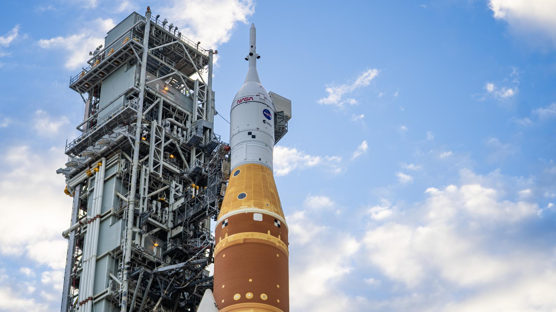 An orange and white rocket is held up by scaffolding with a cloudy blue sky behind it.