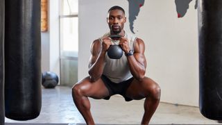 A man in shorts and workout vest in a sparse gym holds a kettlebell to his chest as he does a goblet squat. His knees are bent and pointing outwards, while his chest and head are upright.