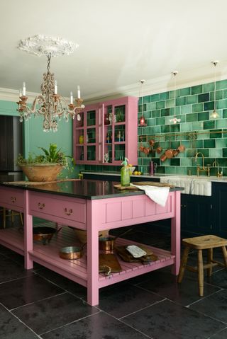 A kitchen with a bright pink kitchen island with a green vase on it, a chandelier above it, and a wooden stool next to it, with dark tile flooring