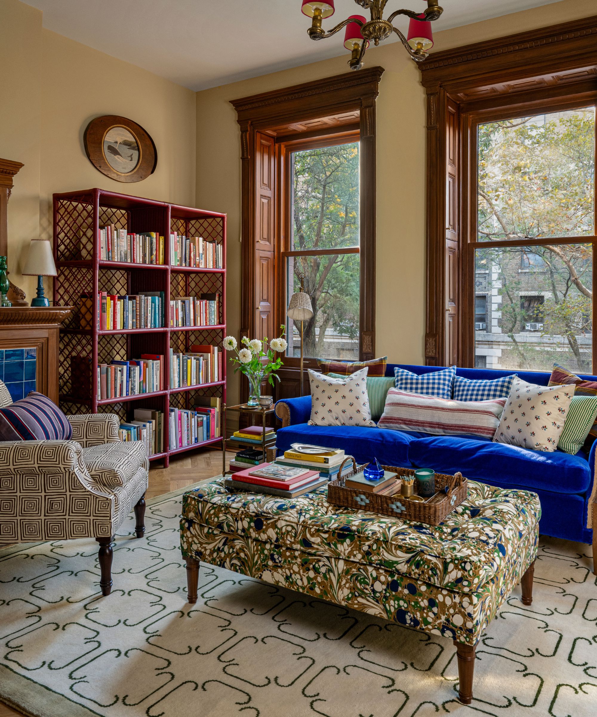 a brownstone home library with yellow walls and tall original wooden casement window, a blue velvet couch and a bookcase