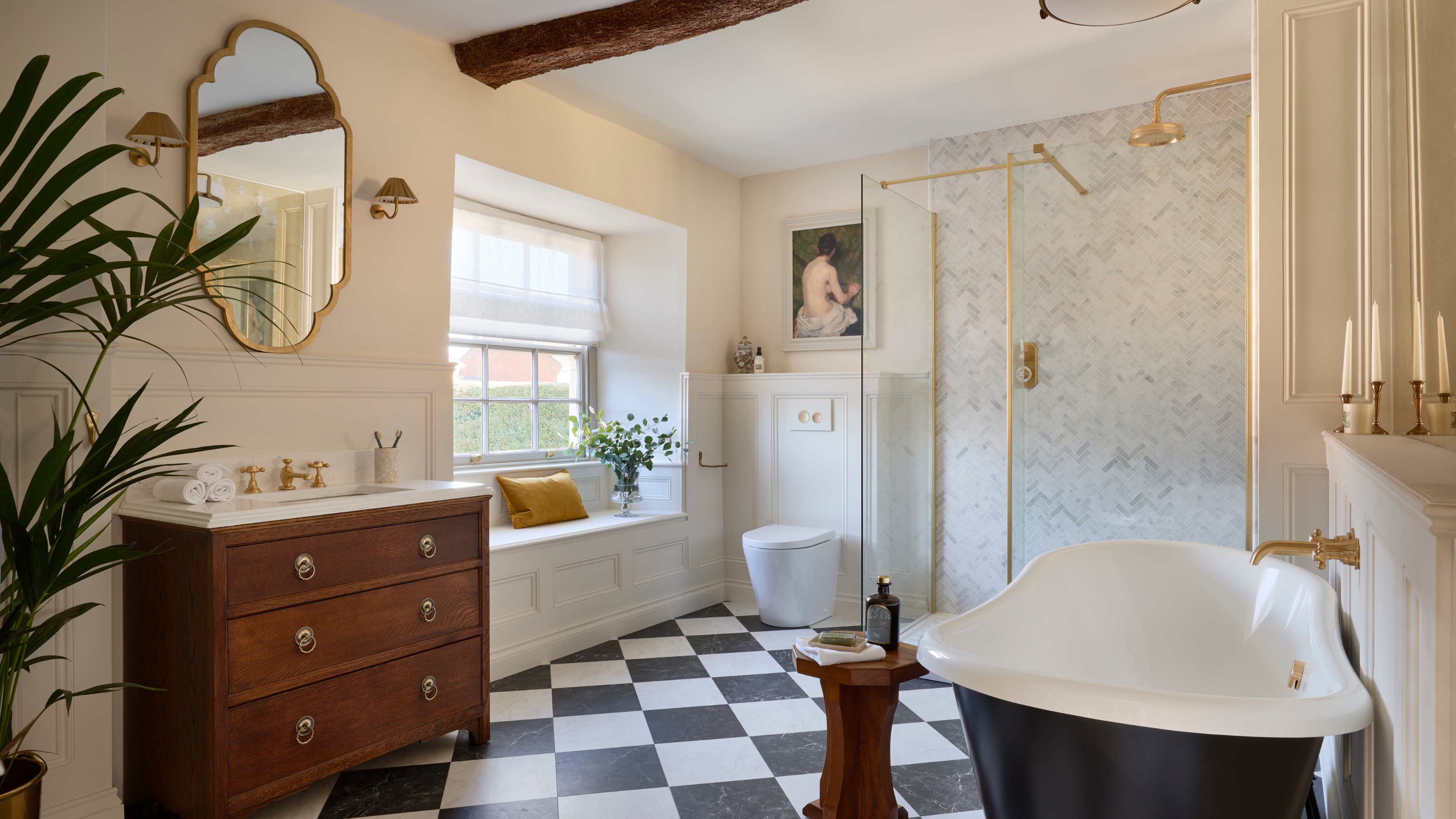 A classical traditional spacious bathroom with chequerboard floor tile, a black freestanding bath, dark wood vanity drawers and brushed brass throughout