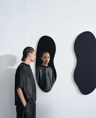 A model dressed in black whose face reflects in a lacquered wood mirror with organic shape.