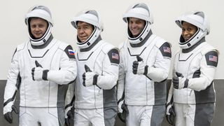four astronauts with white spacesuits and helmets giving a thumbs up while standing in a row