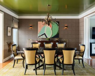 Dining room with vivid green painted ceiling, white ceiling trim, brown patterned wallpaper, oval black dining table with eight upholstered dining chairs in yellow striped fabric, abstract wall art, two wall lights, yellow patterned rug, wooden flooring