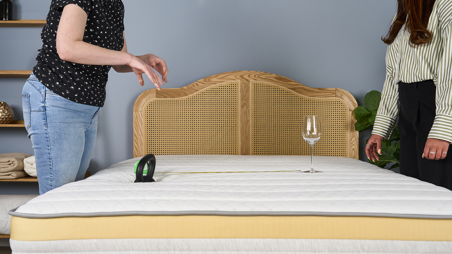 Lead tester Ruth Jones stands on one side of the Helix Dawn mattress while Sleep Staff Writer Eve Davies stands on the other to conduct a motion isolation test. A wine glass is balanced on one side of the mattress, with a measuring tape indicating a distance of 25 inches. At the other end of the tap, a 10lb weight drops onto the mattress. This test helps us to establish the level of motion isolation in the Helix Dawn mattress.