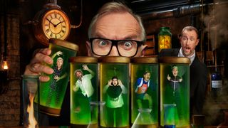 A stylised picture of the Taskmaster season 19 cast, portraying Greg Davies as a giant mad scientist who has trapped each of the contestants in containers full of green liquid. He is holding the container with Rosie Ramsey in it in his right hand (the left hand as we look at the image), and the other containers are holding Jason Mantzoukas, Fatiha El-Ghorri, Mathew Bayton and Stevie Martin. In the background of the laboratory, Alex Horne looks on in alarm.