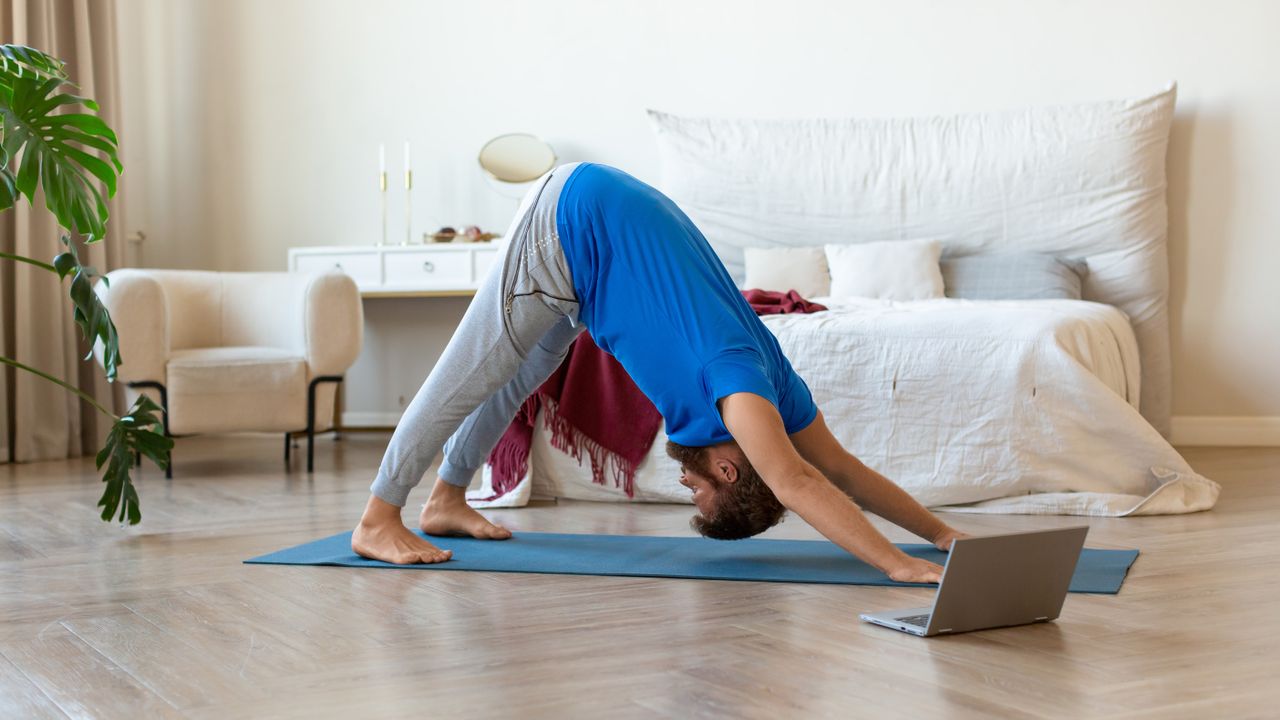 A man performs a downward dog pose in a bedroom on a yoga mat. He is facing downward, with his hands and feet on the mat and his hips held high, so his body forms and upside down V shape. Behind him we see a pristine bed, white arms chair and dressing table.