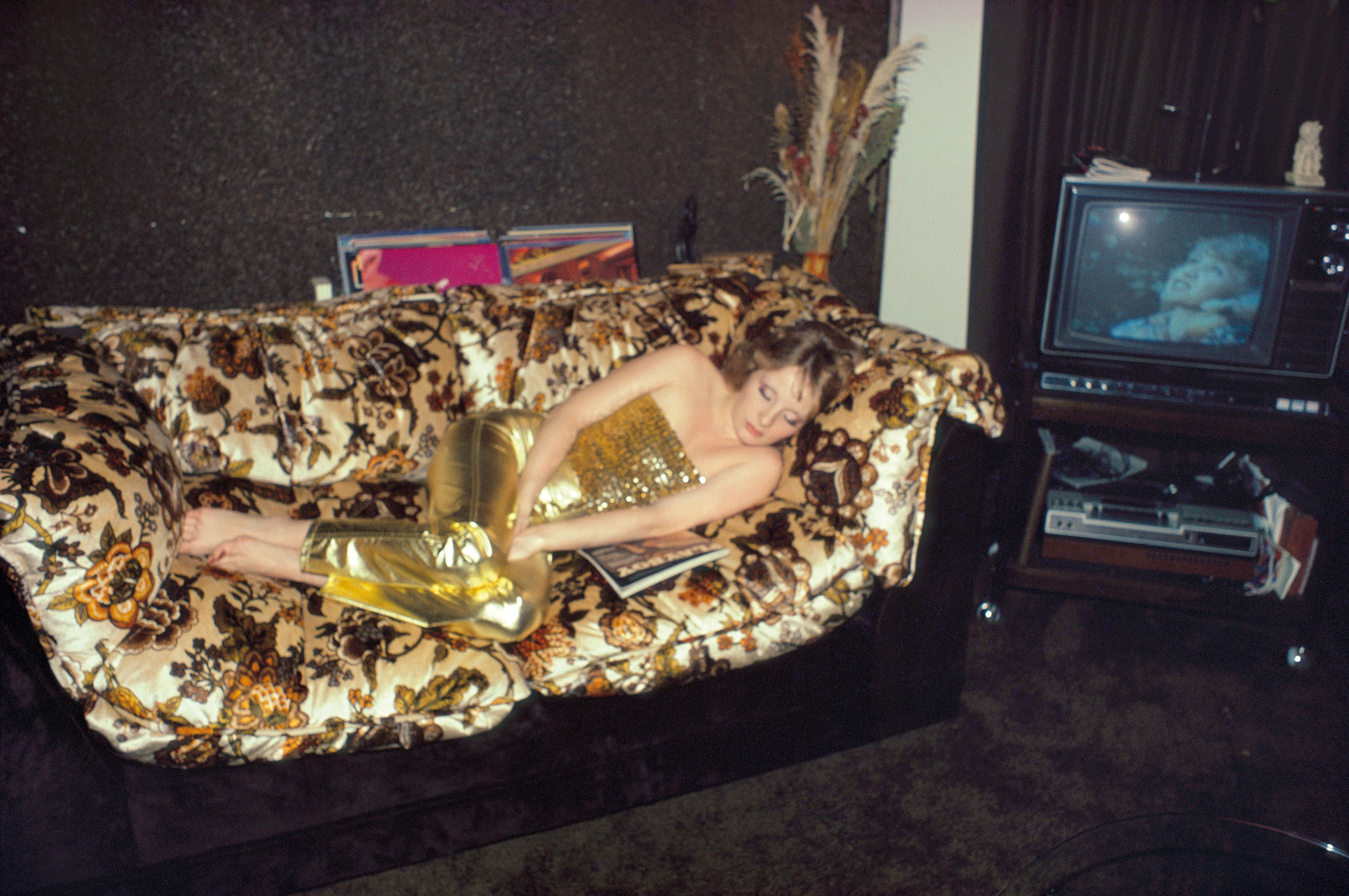 A woman in a shimmering gold outfit lies asleep on a floral-patterned sofa in a room with a vintage television playing in the background.