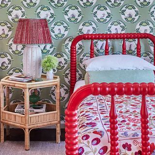 A bedroom with a single red bed with a bobbin frame set against a floral wallpaper