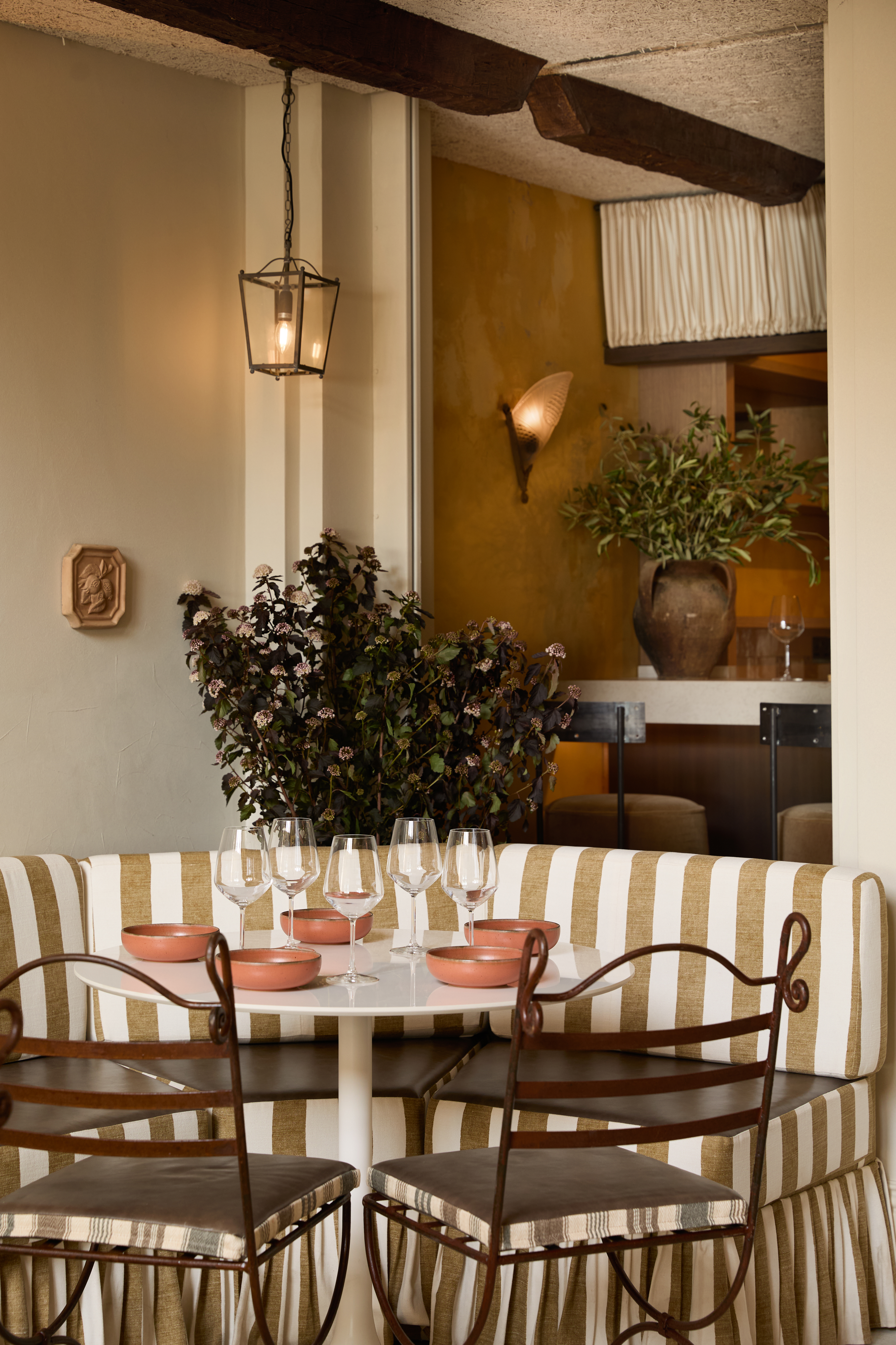 A warmly furnished restaurant with butter and ochre plaster walls, wrought iron chairs with neutral cushions, statement ceilings in brick or plaster relif, and stripy furniture paired with plants.