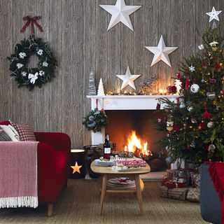 a cosy country living room with wood panel wallpaper, a roaring fire with a white mantle, a red armchair, a decorated christmas tree with presents underneath and a wreath and white star decorations on the wall