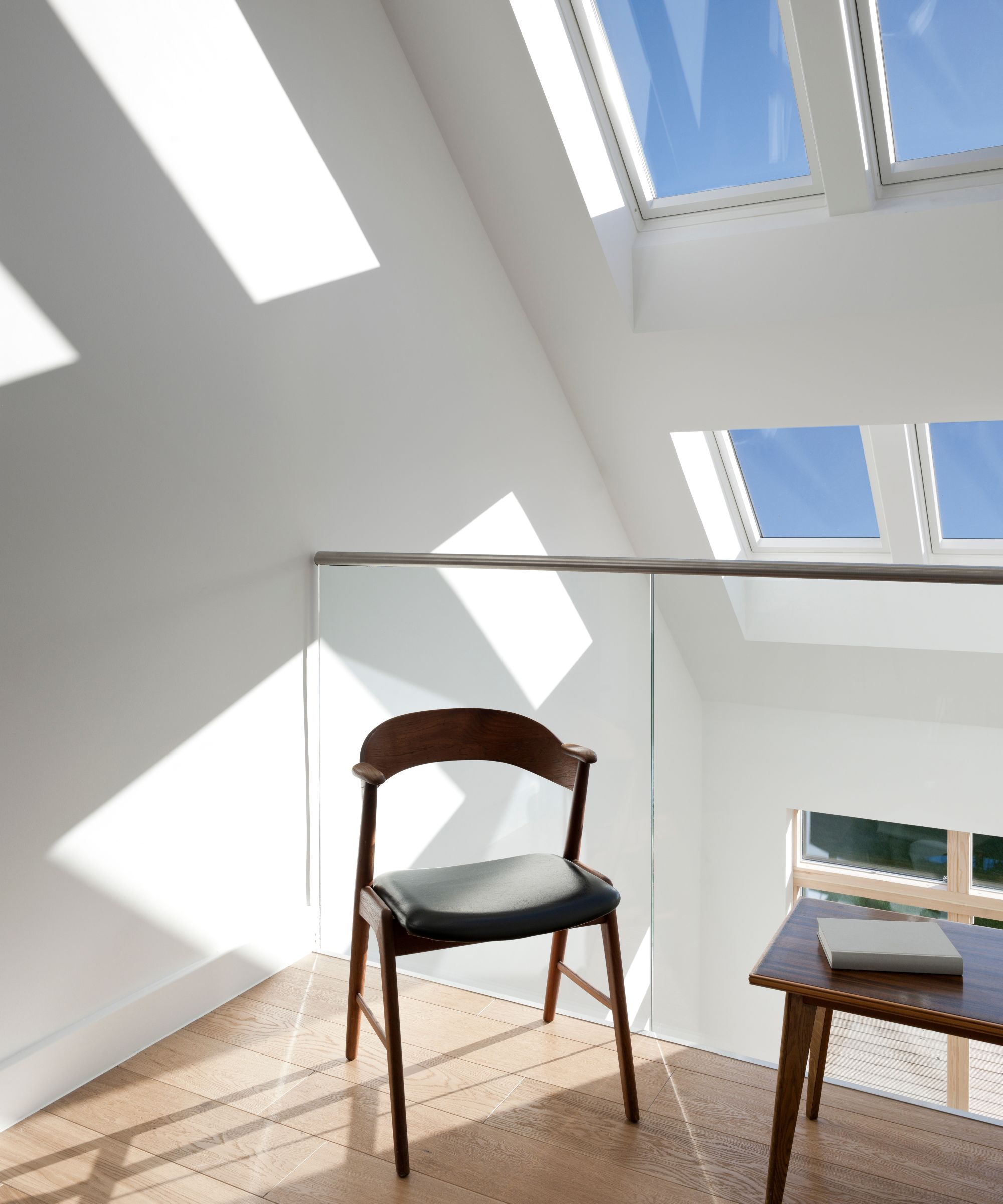 Bright, minimalist landing area flooded with sunlight from large roof windows. A dark wood chair and small side table create a peaceful nook with clean lines and natural warmth.