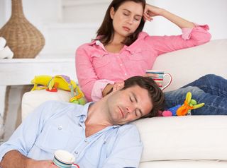 Parents sit in their living room, exhausted, amid children's toys strewn everywhere.