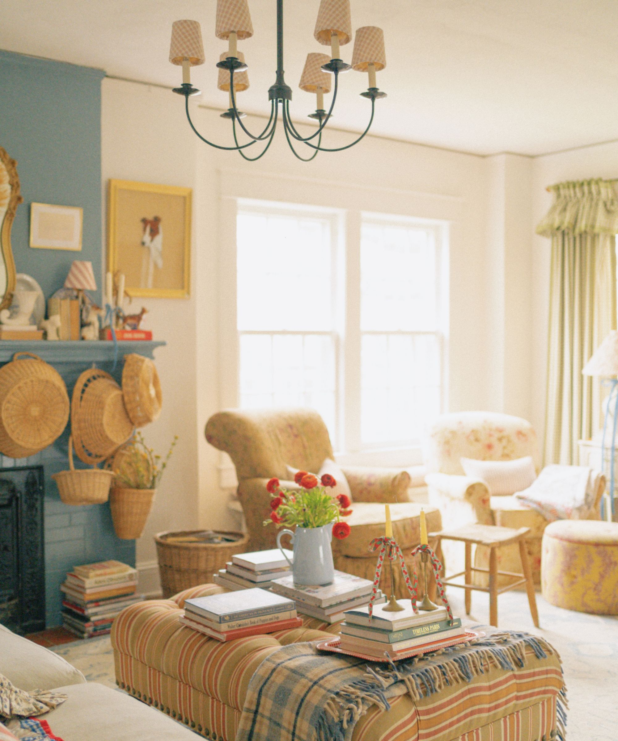 Vintage living room with upholstered ottoman, blue painted fireplace, baskets hanging, floral armchairs, and iron chandelier