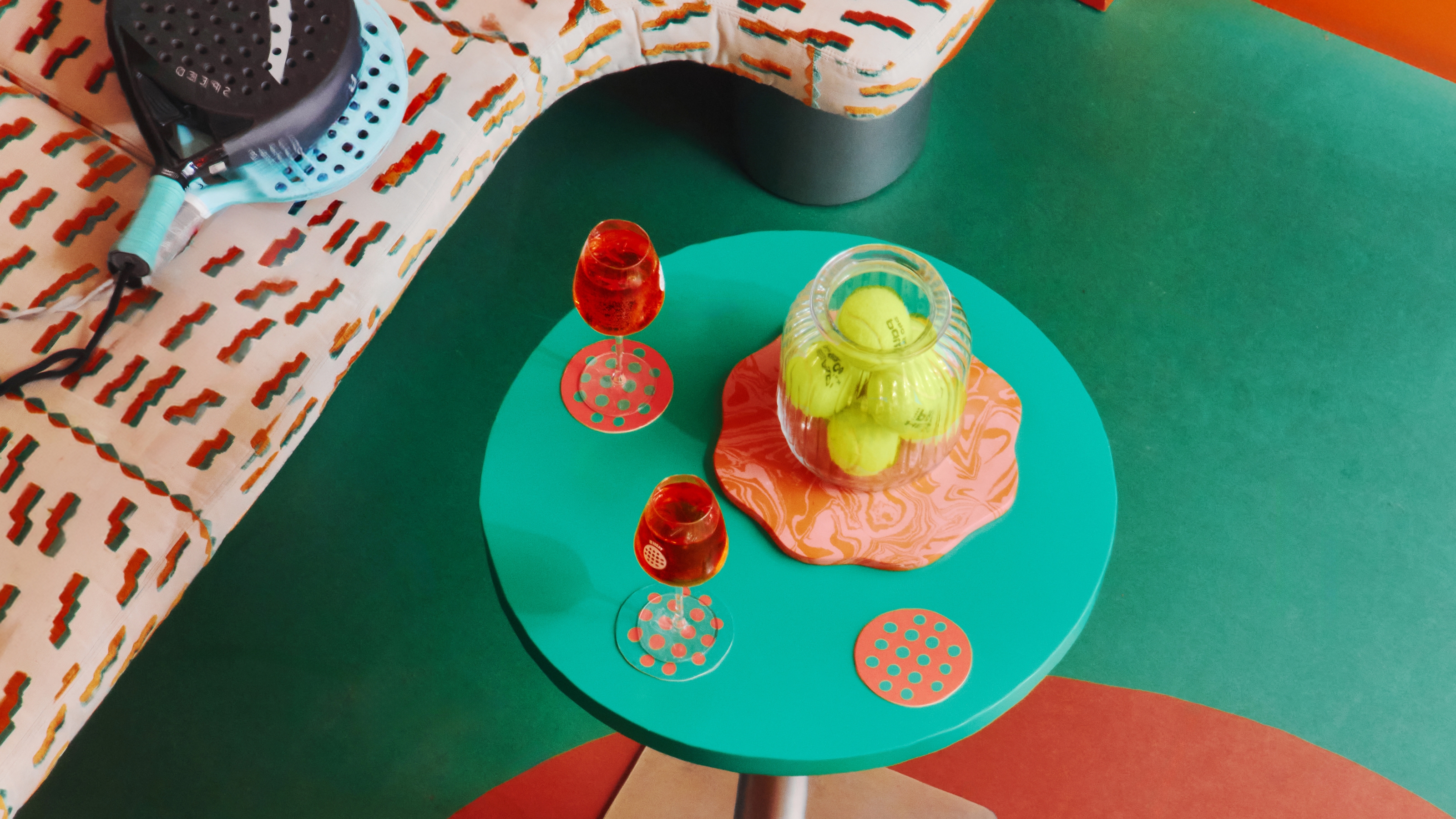 A padel bar's furniture, including an emerald green round table with a chrome base, topped with orange coasters, glasses, and a jar of padel balls, and rackets laying on top of an upholstered cream, orange, and green banquette, captured from above.