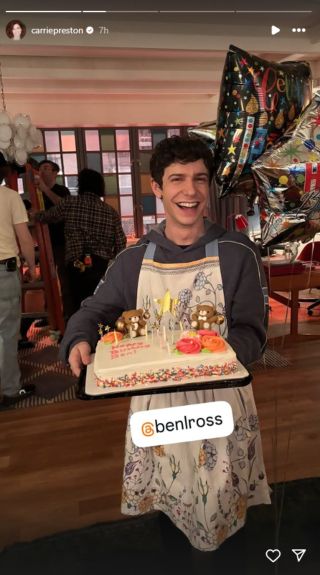 Ben Levi Ross holding a cake and smiling while wearing an apron.