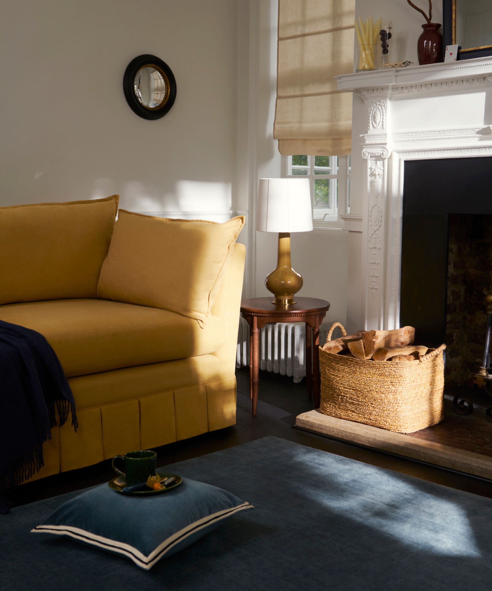 Yellow sofa with pleated skirt with blue rug, fireplace, timber side table with lamp, woven basket filled with firewood, small round mirror on wall, and roman blinds on window