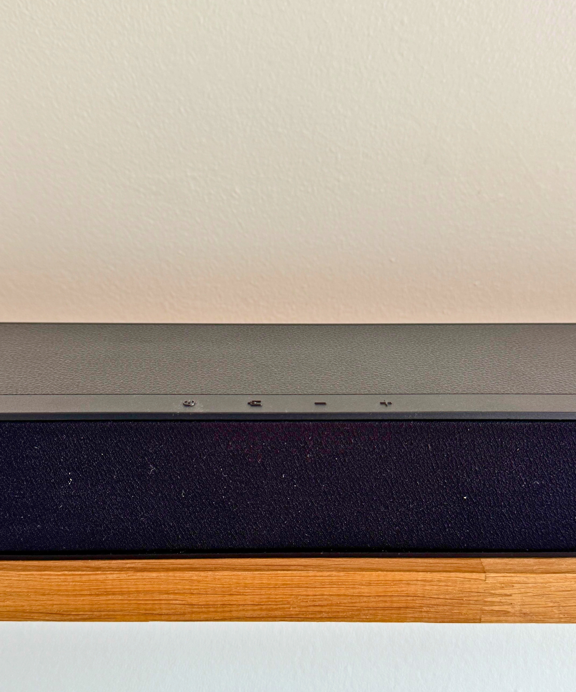 Closeup of Hisense HS2100 soundbar's four control buttons on wooden shelf in front of white wall.