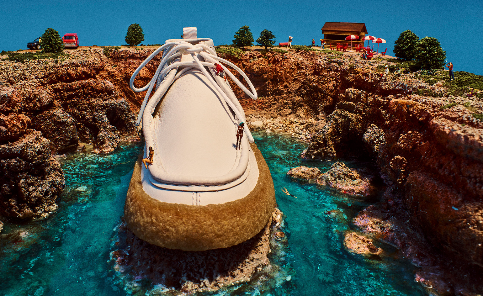 View of a large white and brown shoe with laces surrounded by water and cliffs. On top of the cliffs are miniature people, trees, cars, a wooden cabin, red chairs and benches and red and white parasols. There are also miniature people on the shoe