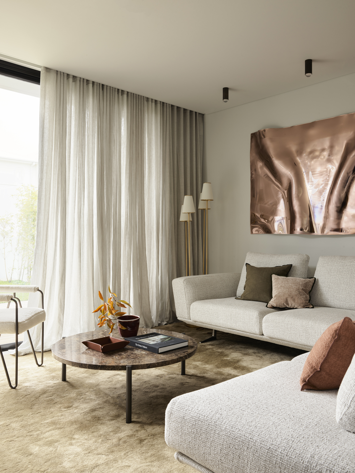 beige living room with glossy chrome artwork, golden carpet, beige sheers, beige sofa, and round stone coffee table styled with books and a vase