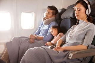 A woman wearing headphones and a man wearing a neck pillow on a flight with a young girl sat in between them