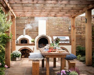 An example of outdoor kitchen ideas showing a wood-fired pizza oven, grill and wooden table and benches