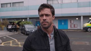 Olly Rix as Flynn Byron outside Holby ED looking serious and wearing a grey polo shirt and a black jacket. 