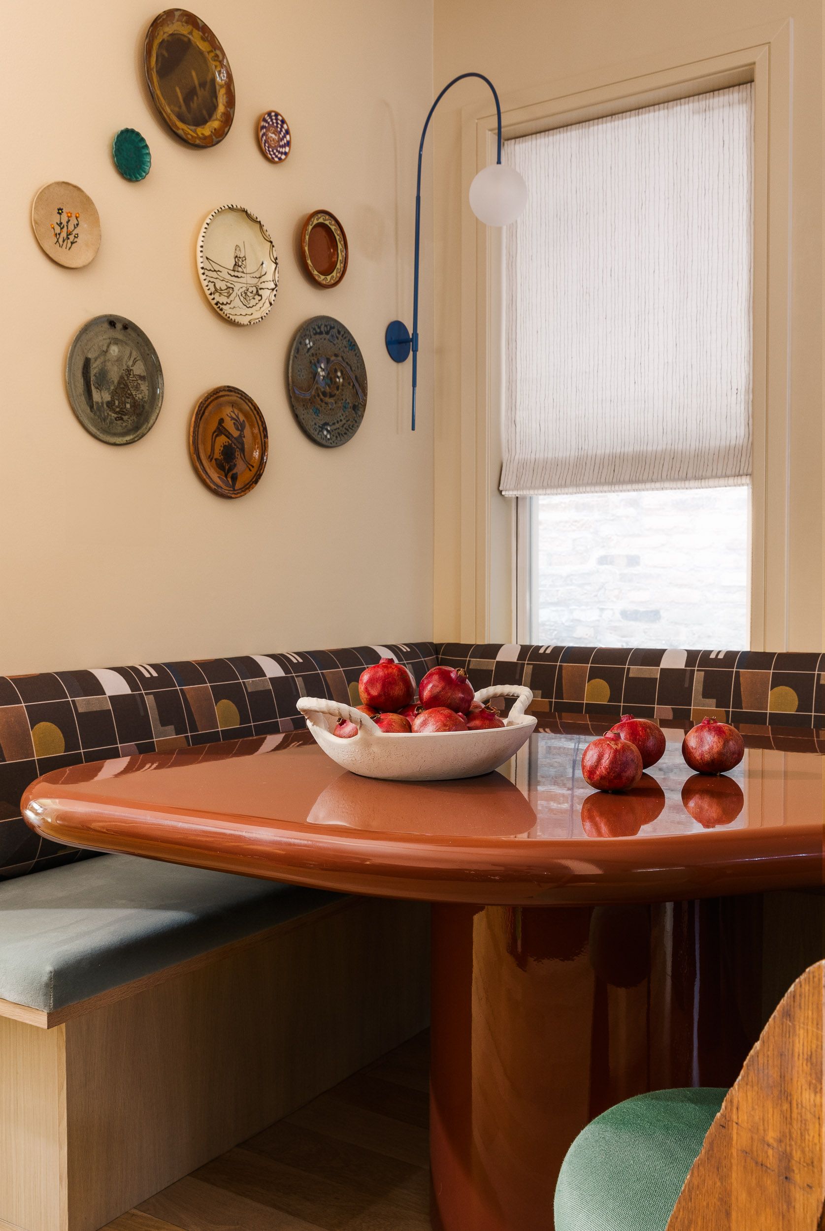Image of a dining corner nook with a patterned bench seat and a red lacquer table. There is a white dish filled with pomegranates and plates hung on the wall. 